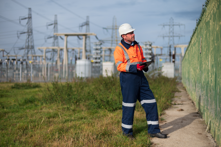 Engineer wearing PPE and holding a tablet inspecting a fence by an electricity substation