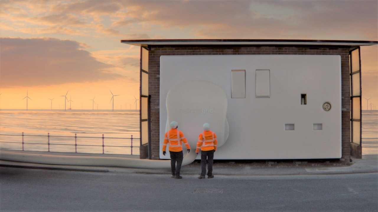 Two persons wearing orange high-vis jackets and hard hats seen from behind standing in front of a giant plug and socket with the sea behind