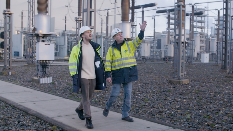 Two people wearing hard hats and high-vis jackets walking with electricity equipment in the background