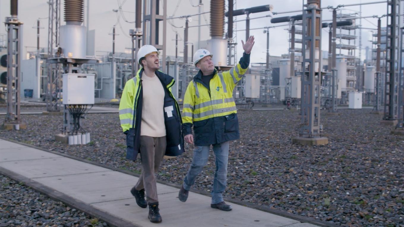 BritNet_JamesStewartAndTommyHoogstrate_1920x1080.jpg Two people wearing hard hats and high-vis jackets walking with electricity equipment in the background