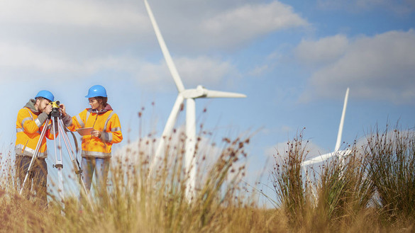 Man and woman standing in grassy field in front of windfarms, wearing hard hats and high-vis jackets with a survey camera