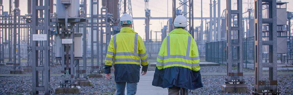 Two people wearing helmets and high-vis jackets walking through an electricity substation