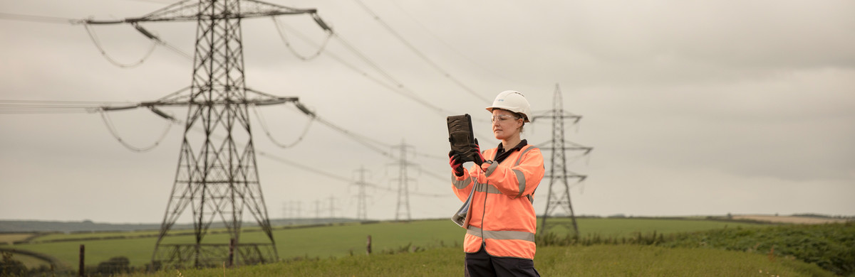 Woman wearing hard hat and orange high-vis jacket looking at tablet in green field with pylons