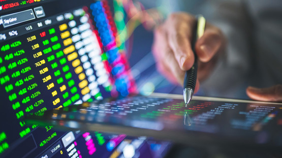 Hand holding a pen working on a smart-pad with stock market trading screens to the side